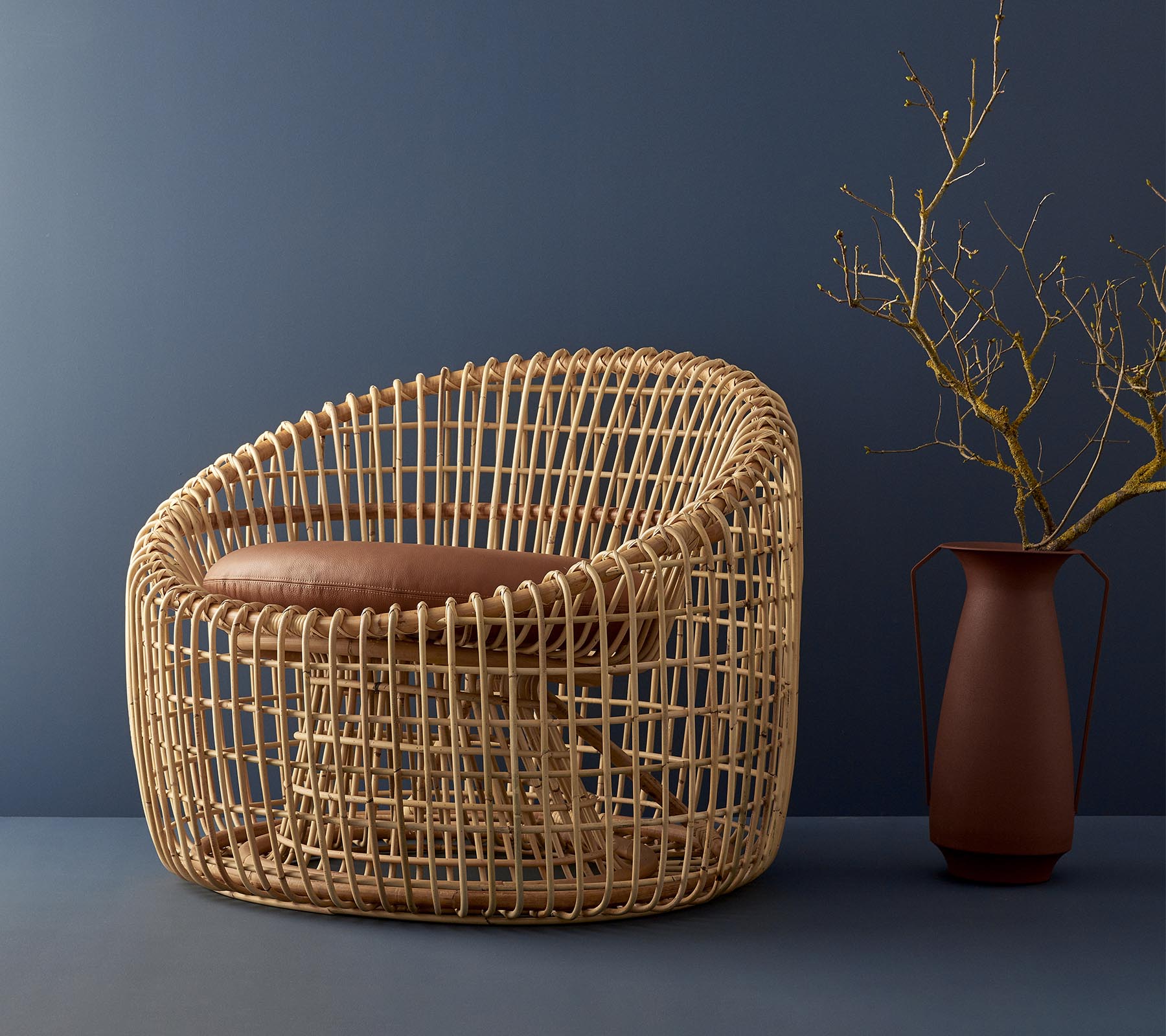 Modern korgstol med rundad design, med en stoppad sits och kompletterad av en enkel vas med en kvist.