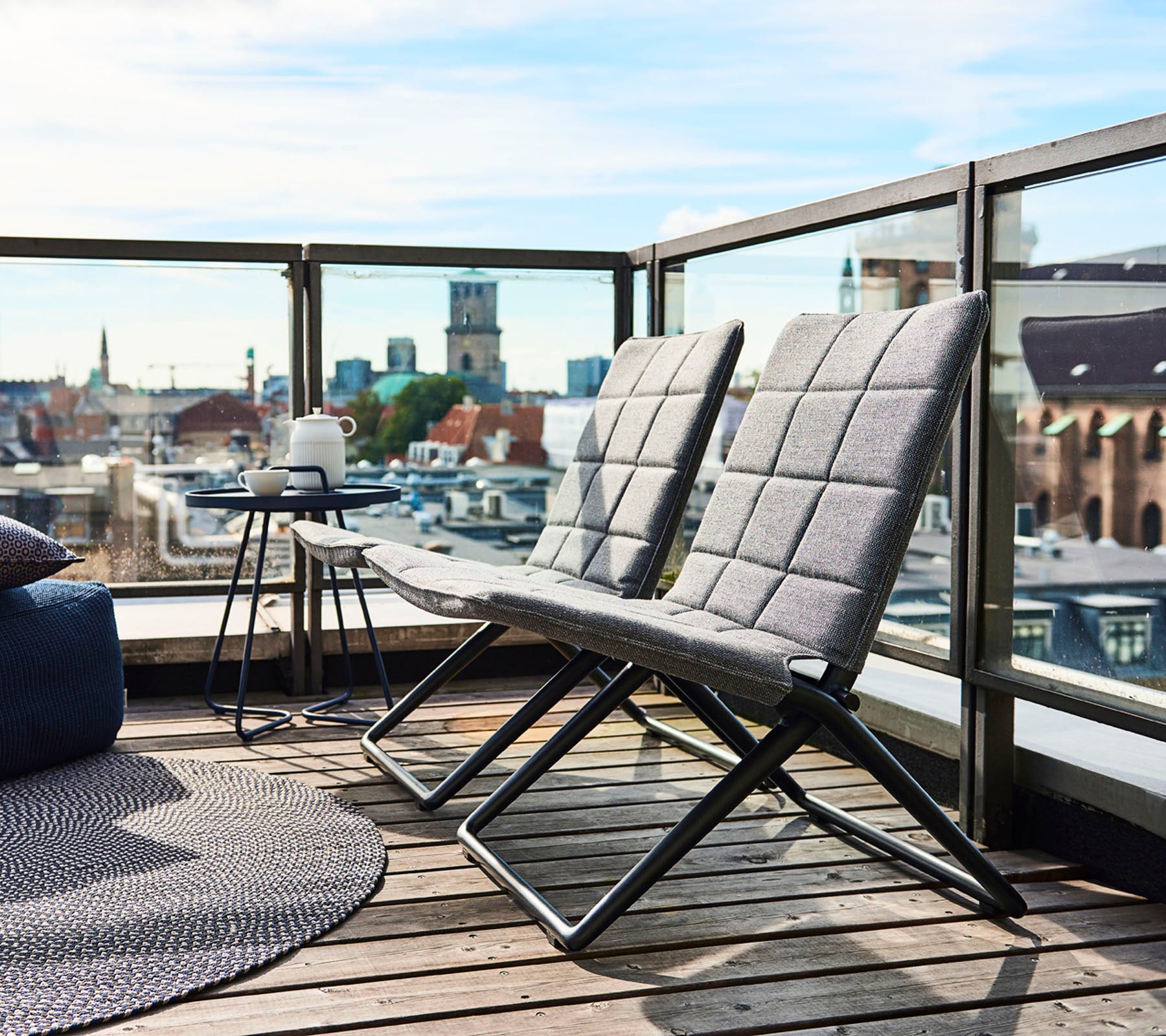 Två moderna loungefåtöljer med slanka ramar och texturerade kuddar, i sällskap av ett litet sidobord och utsikt över stadens landskap i bakgrunden.