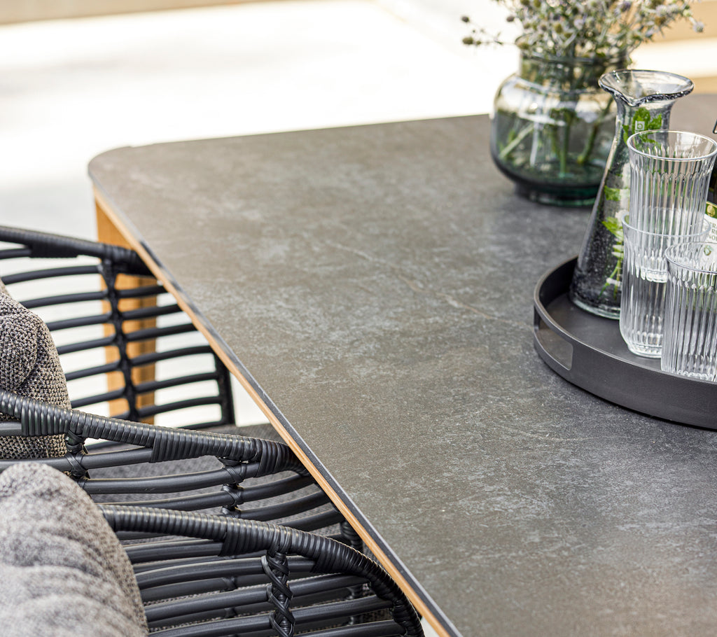 En modern middagsuppsättning med ett slätt bord, en stiliga korgstol, glas på ett fat och en dekorativ vas med grönska.