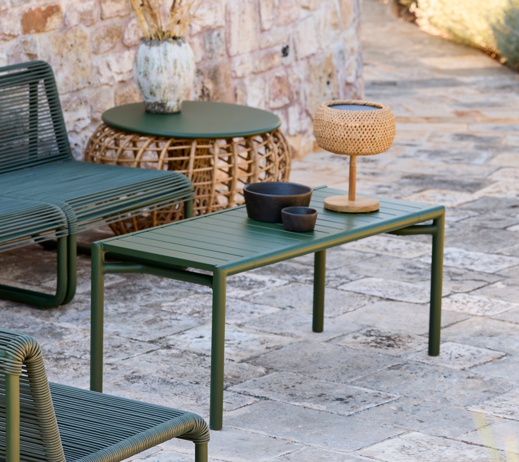 Grön bord med svart skål, brun vas och vävd lampa i en modern utomhusloungedel.