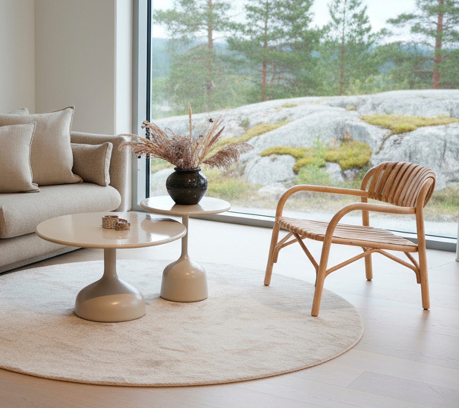 Modern lounge med en soffa, runt bord, svart vas och rottingstol nära ett stort fönster.