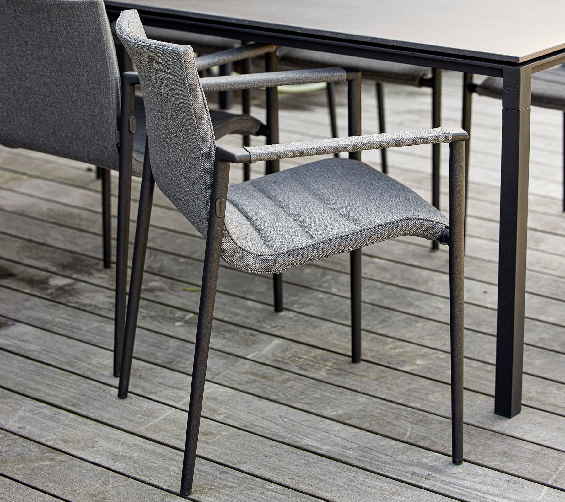 Stilfull grå stol med armstöd placerad bredvid ett elegant svart bord på en trövyta. Ideal för moderna utomhusmiljöer.