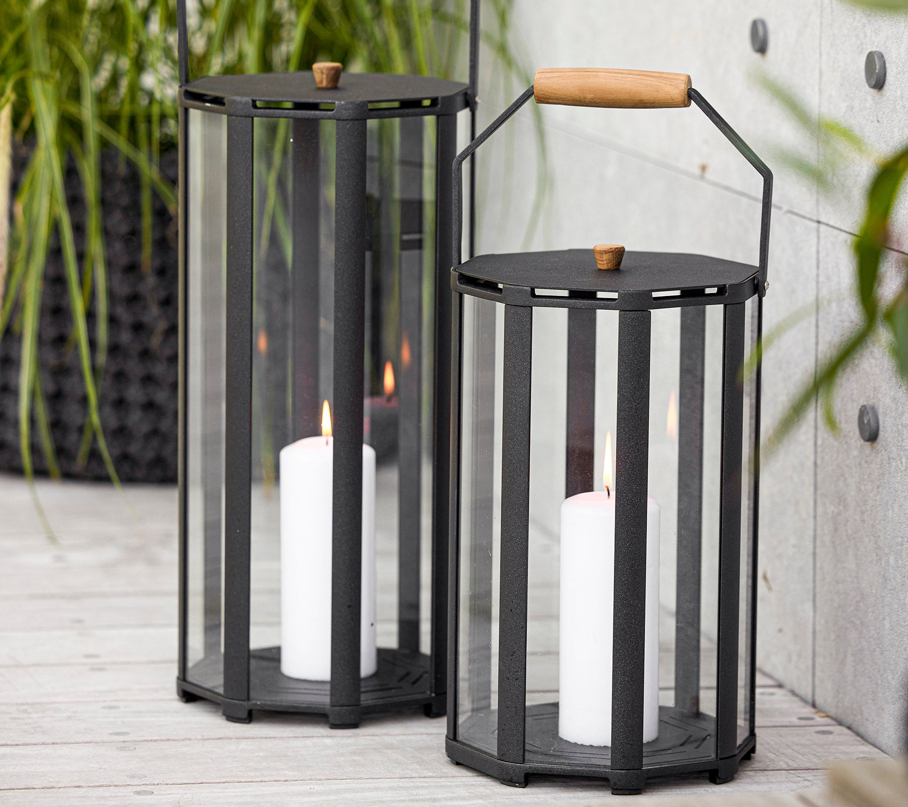 Två stiliga lyktor med modern design, som har glaspaneler och ett trähandtag, som håller vita ljus inuti.