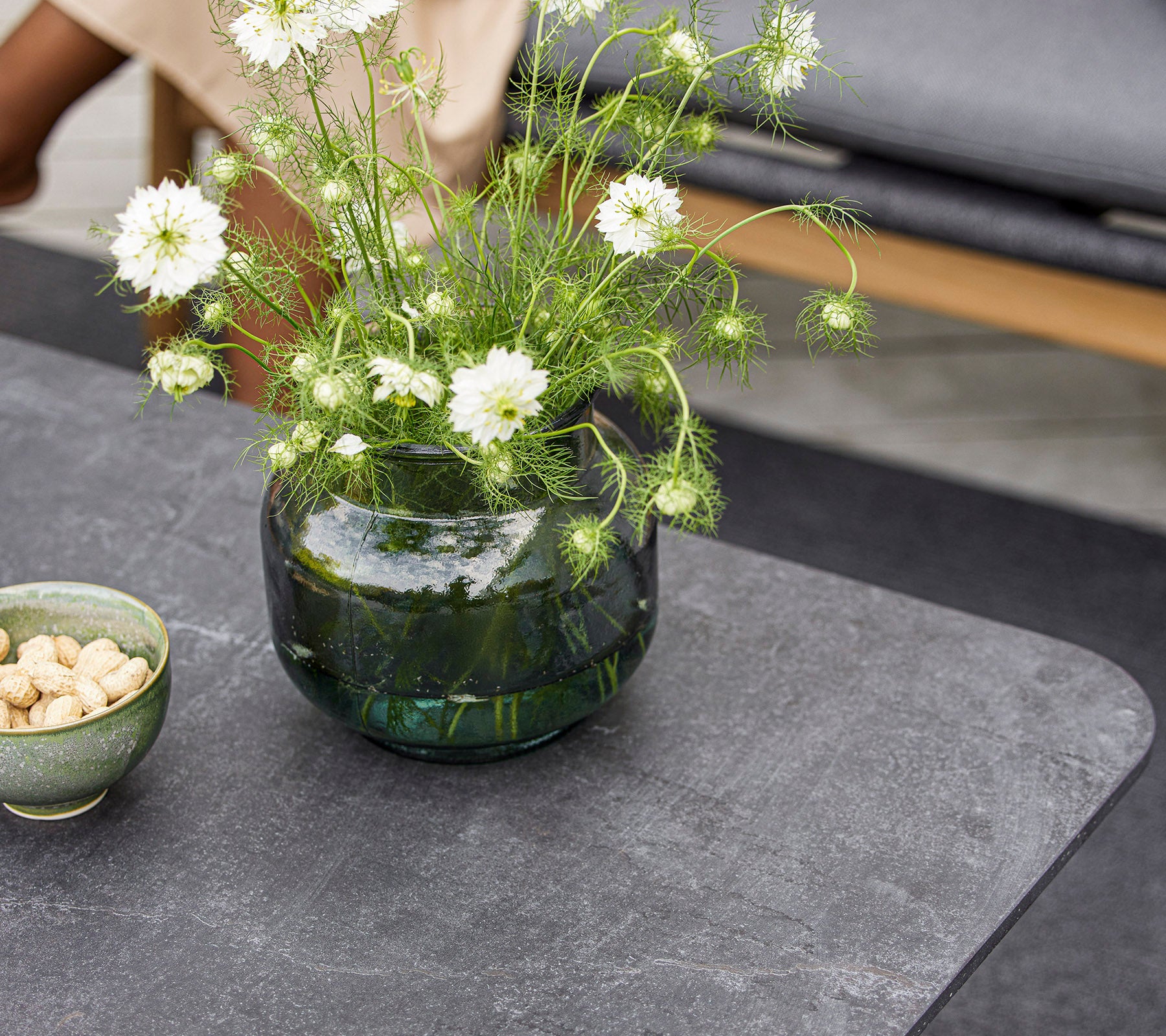 Ett modernt bord med en rund glasvas med vita blommor och en liten skål med nötter. Ytan är slät och texturerad.