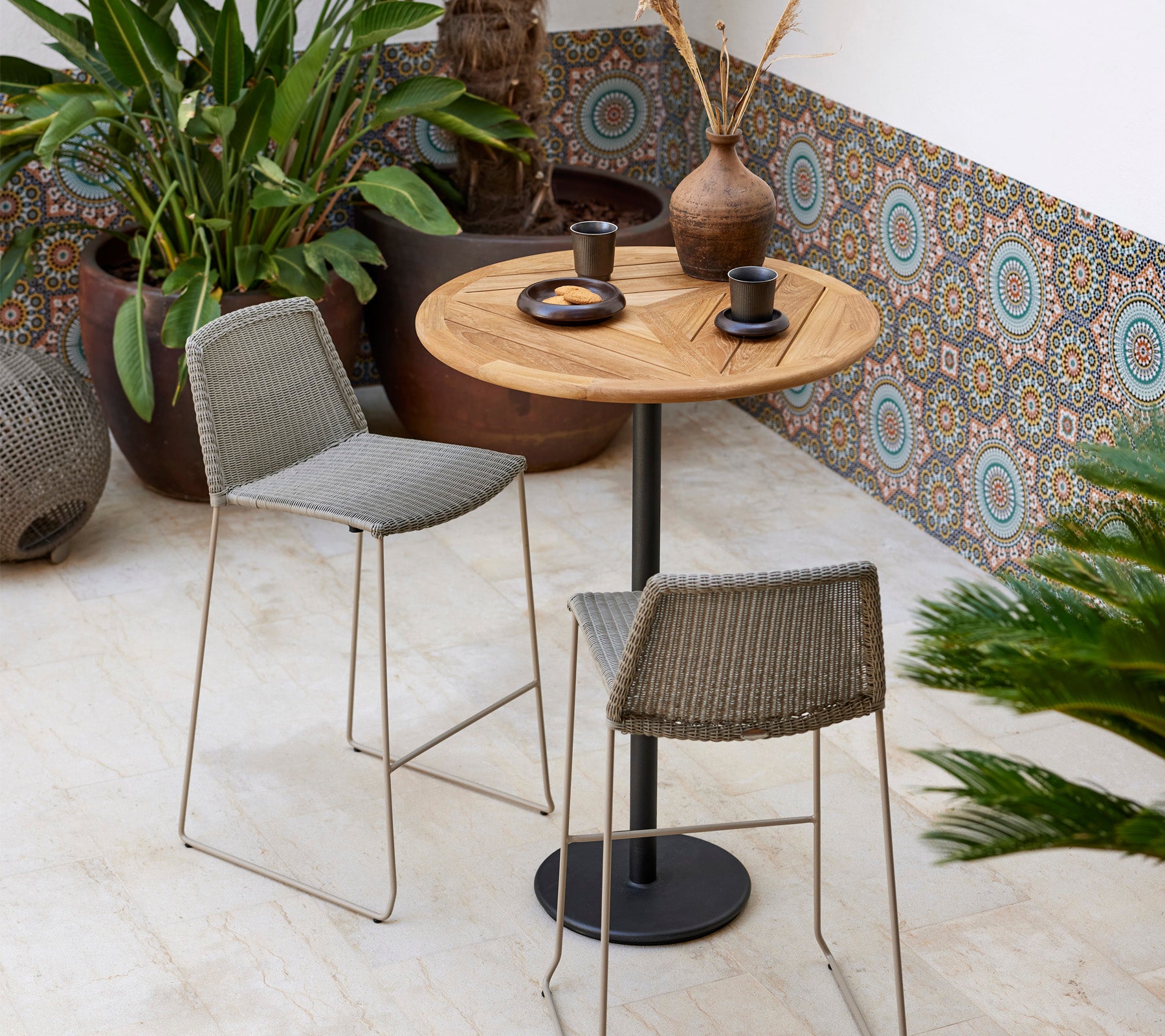 Ett stiligt bistroset med ett runt trädgårdsbord och två vävda pallar, kompletterat med dekorativa växter och modern porslin.