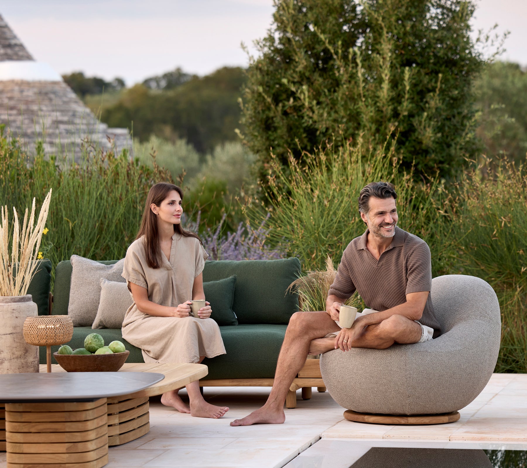 Modern lounge med grön soffa, grå rund stol och brun bord omgiven av grönska.