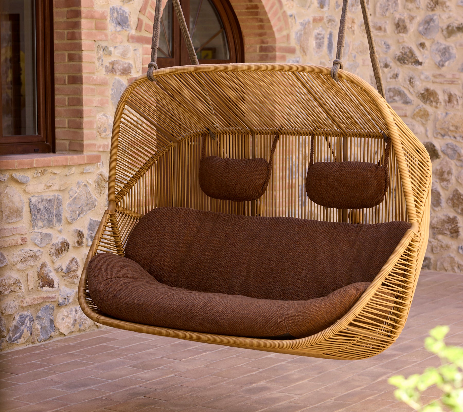 Hanging 2-seater sofa with a woven rattan frame and brown cushions, offering a cozy spot for relaxation.