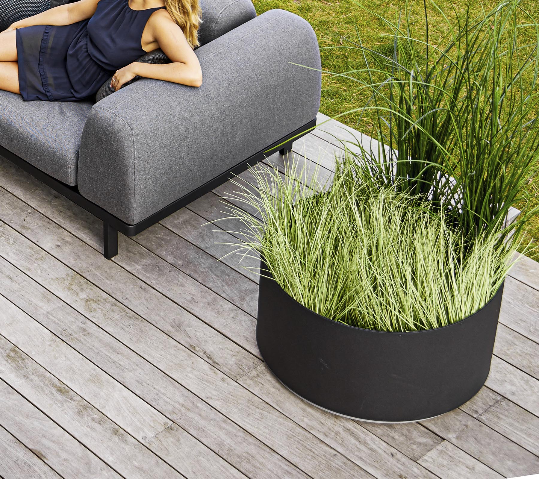 En stilren grå soffa bredvid en rund svart planter fylld med högt grönt gräs, placerad på trädeck.
