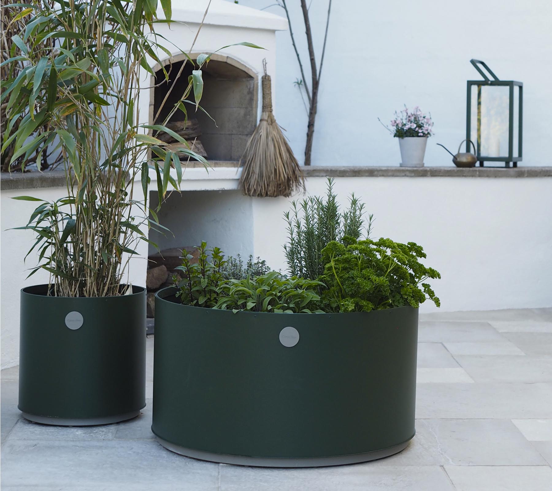 Två runda planter i mörkgrön, den ena med höga växter och den andra fylld med olika örter, står på en ljus stenyta.
