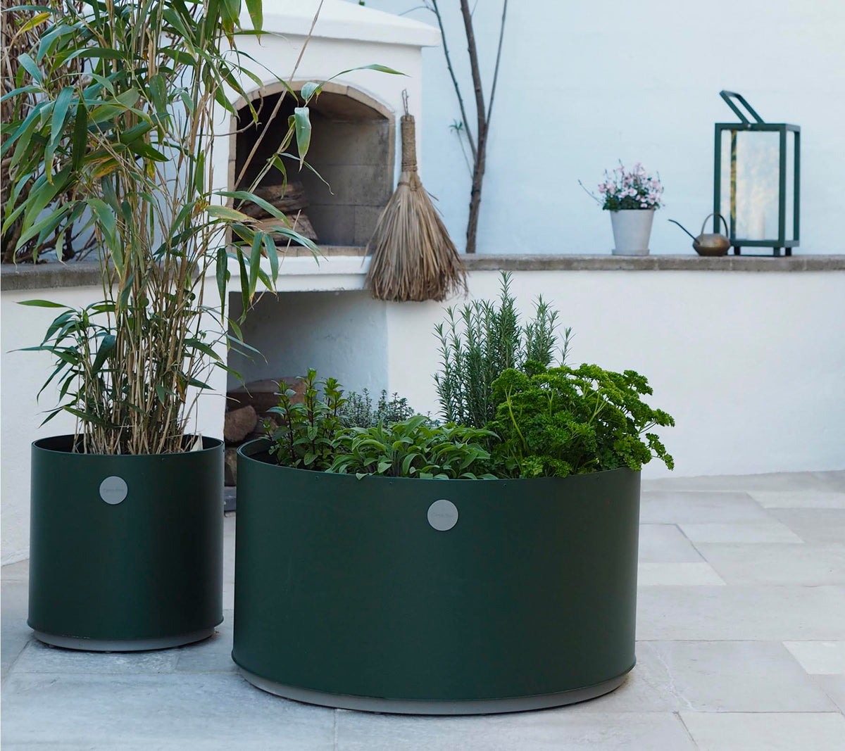 Två runda planter i mörkgrön, den ena med höga växter och den andra fylld med olika örter, står på en ljus stenyta.
