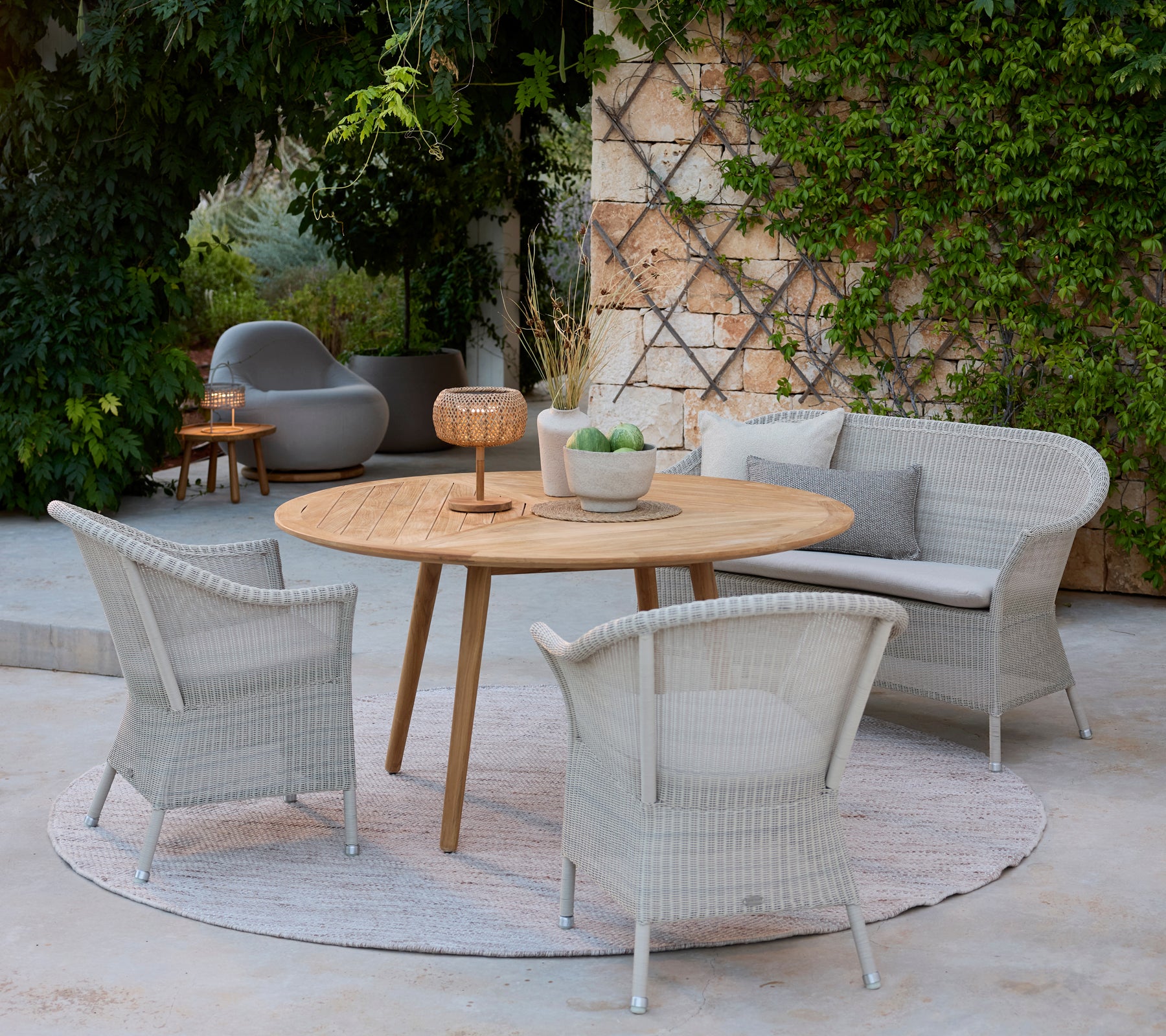 Modern loungeutrymme med grå stolar, ett runt träbord och en grå soffa omgiven av grönska.