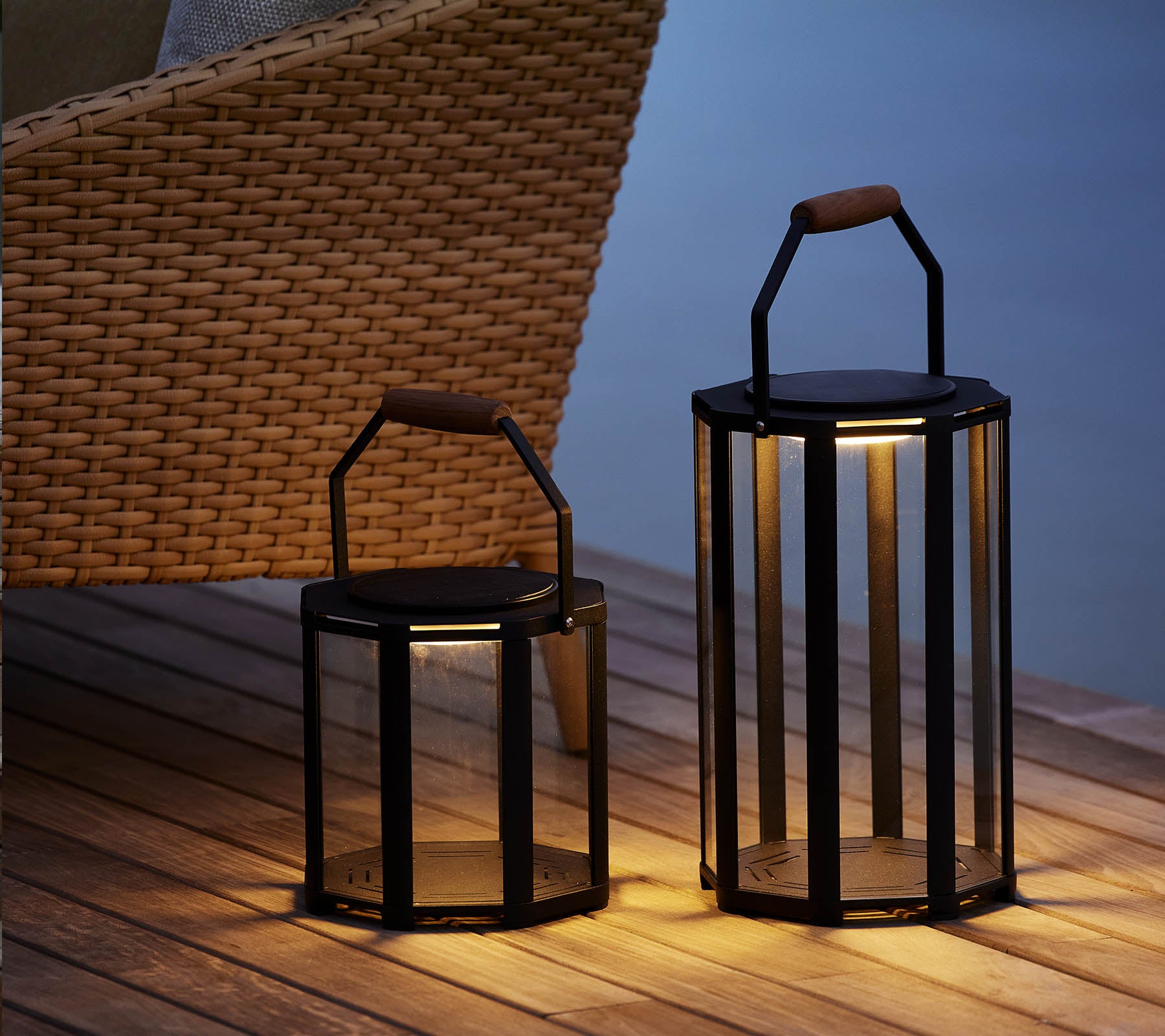 Två stiliga Lightluxe-lampor med glaspaneler och handtag, som ger ett varmt sken, placerade bredvid en vävd stol.