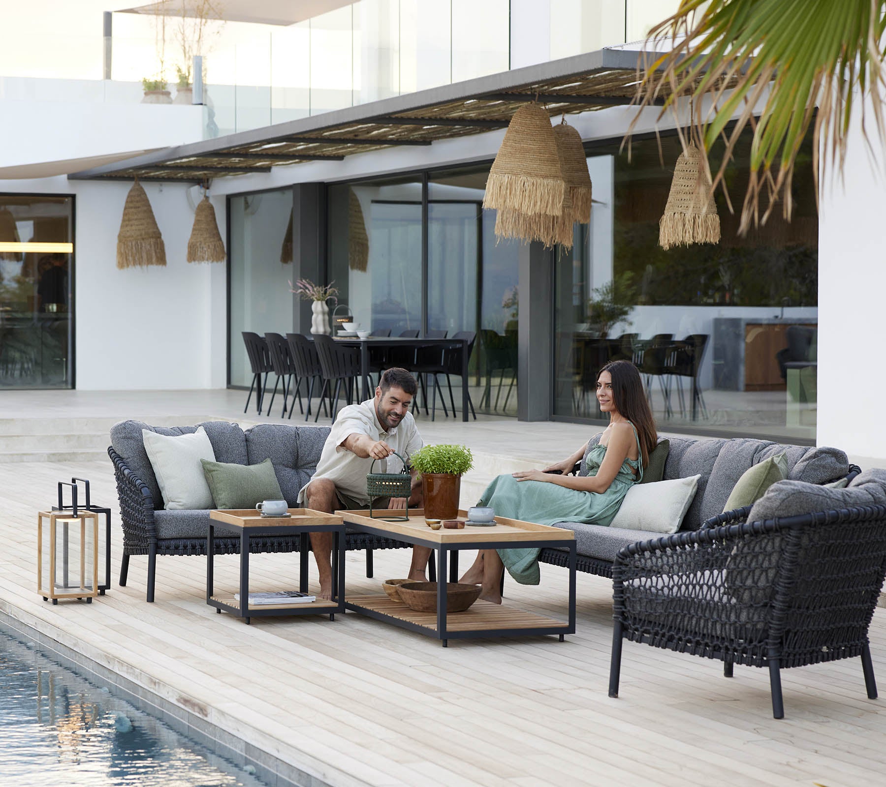 Par njuter av fritid på en mysig loungesoffa, omgiven av gröna växter och stilfull dekoration, med en lugn swimmingpool i närheten.