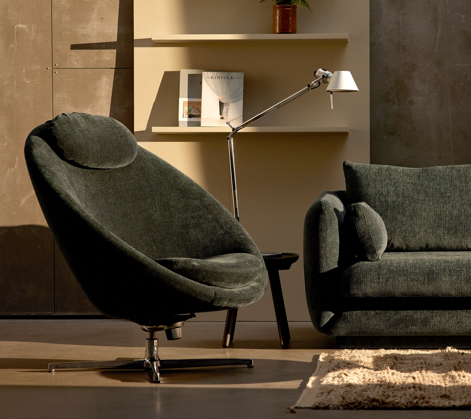 Pace-stol i mörkgrön chenille bredvid en Bloom-soffa, kompletterad av en stilren lampa och minimalistisk hylldekoration.