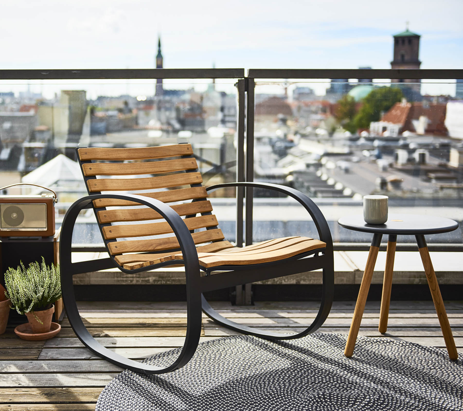 En stilren gungstol bredvid ett litet sidobord, med en panoramautsikt över staden, prydd med en växt och en matta.