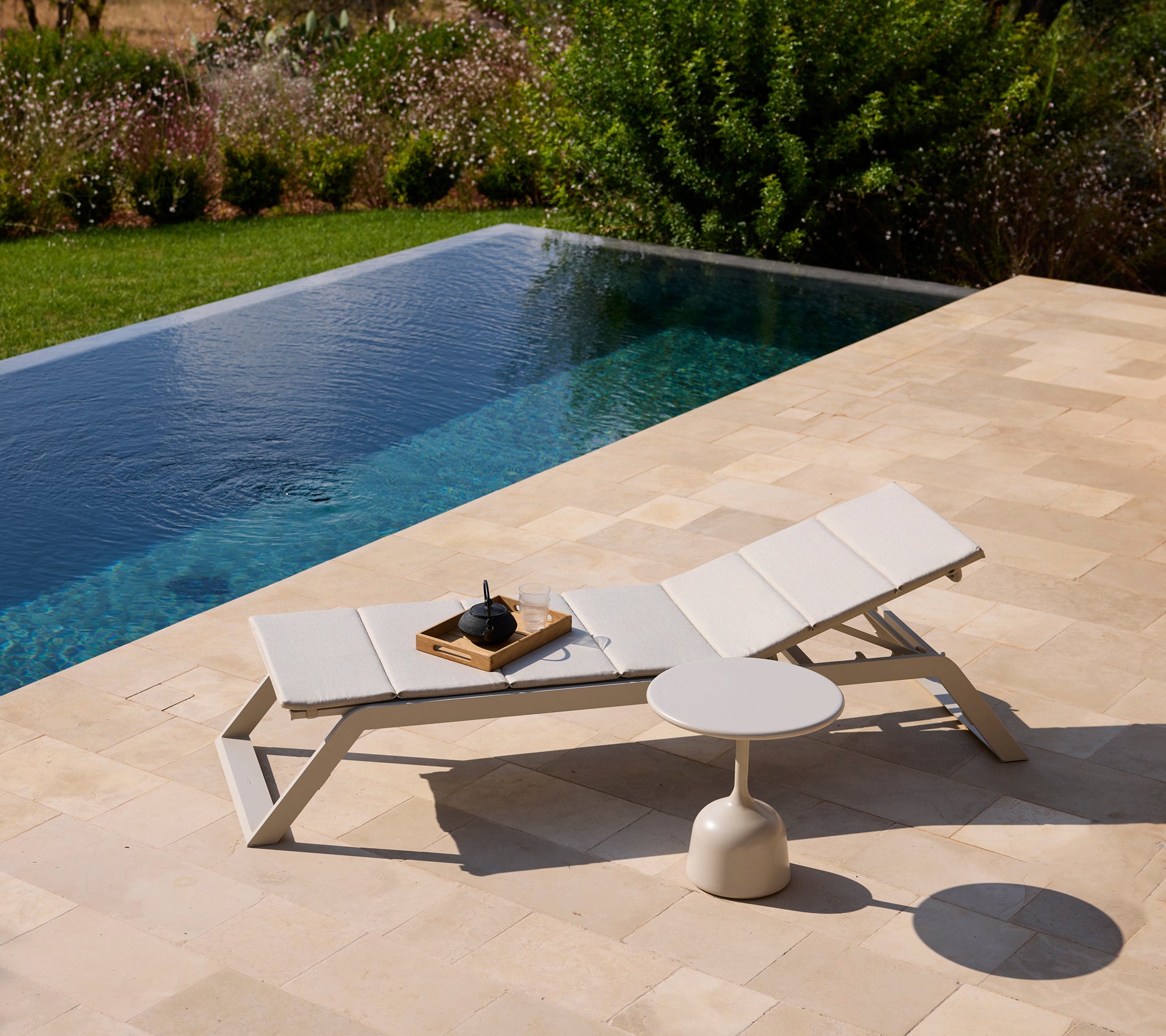Modern vit loungestol och sidobord vid en pool omgiven av grönska.