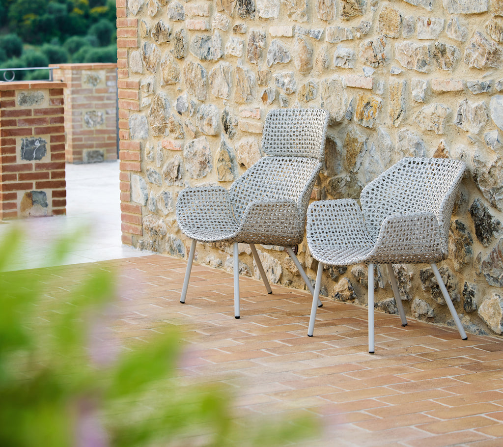 Två Vibe högback loungestolar i mörk sand, stiligt designade, placerade mot en texturerad stenvägg som ger en varm, inbjudande atmosfär.