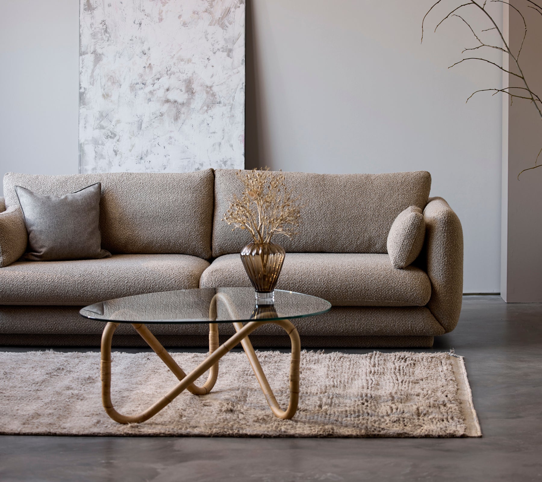 En modern beige soffa med ett glas soffbord och en dekorativ vas i en minimalistisk lounge-miljö.