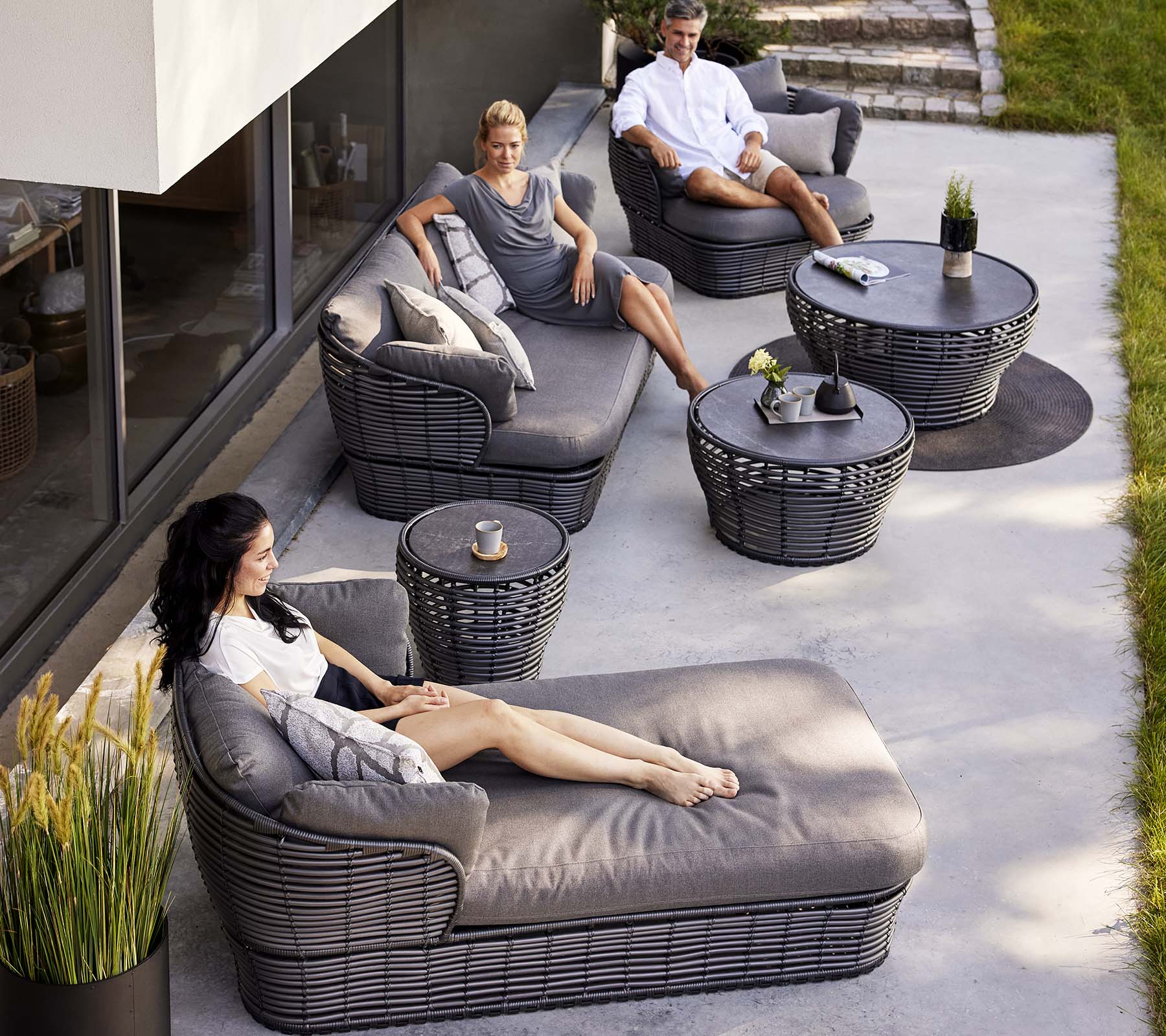 Moderna loungeområde med en korgsäng, en 3-sitssoffa och loungestolar, kompletterat med runda bord och dekorativa växter.