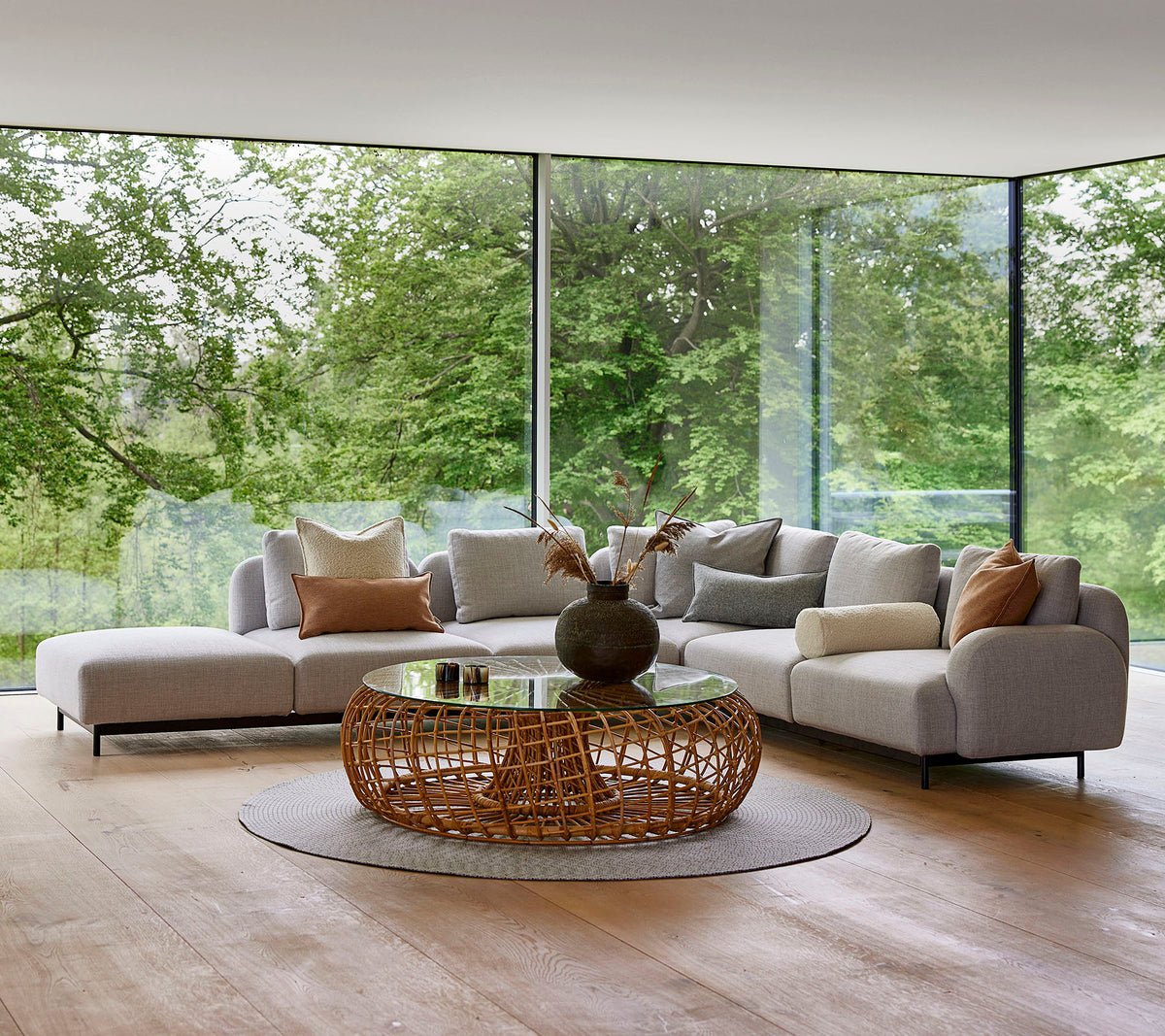 Moderna loungutrymmen med en modulsoffa med dekorativa kuddar, ett runt vävt soffbord och stora glasfönster som visar grönska.