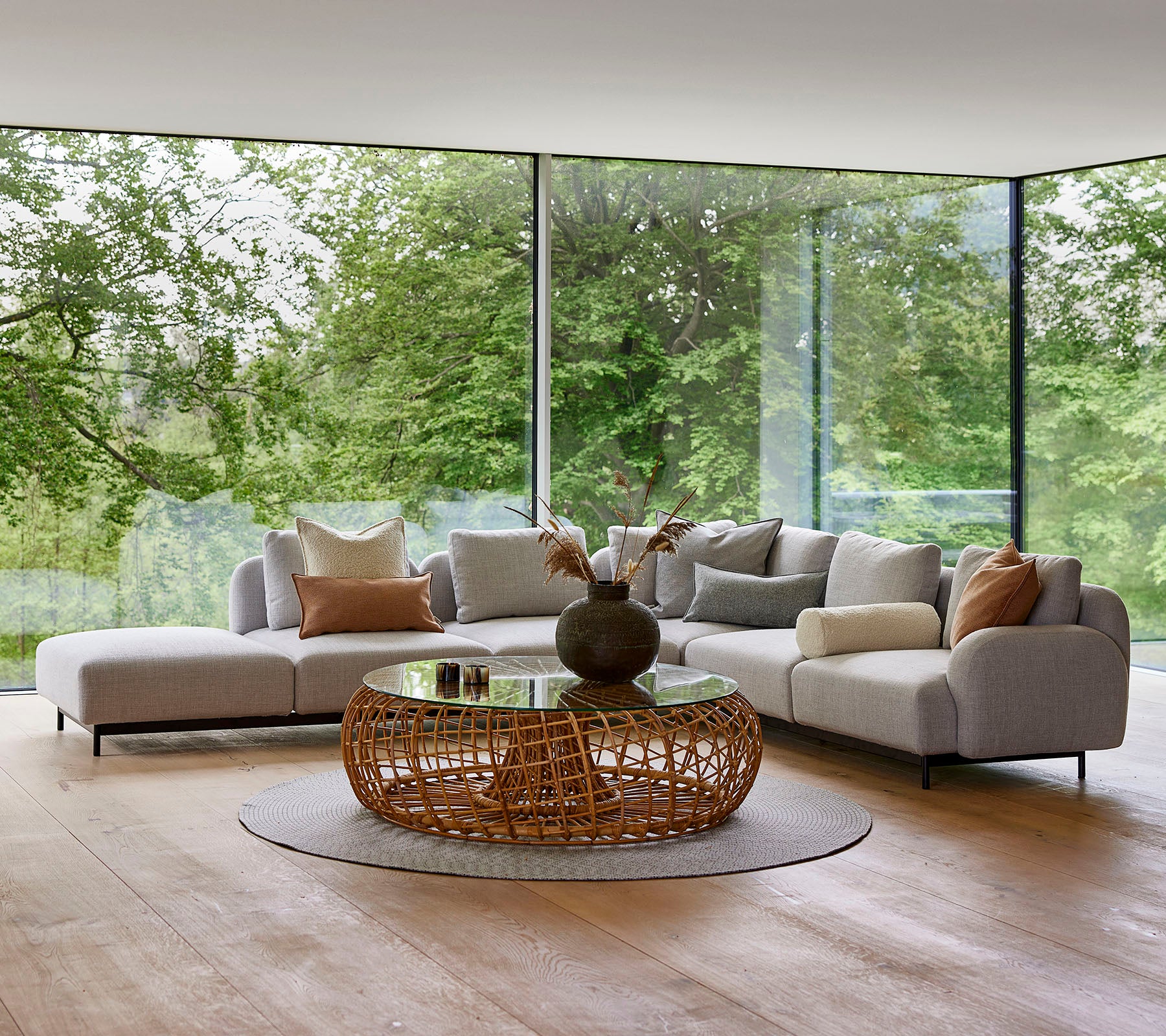 Moderna loungutrymmen med en modulsoffa med dekorativa kuddar, ett runt vävt soffbord och stora glasfönster som visar grönska.